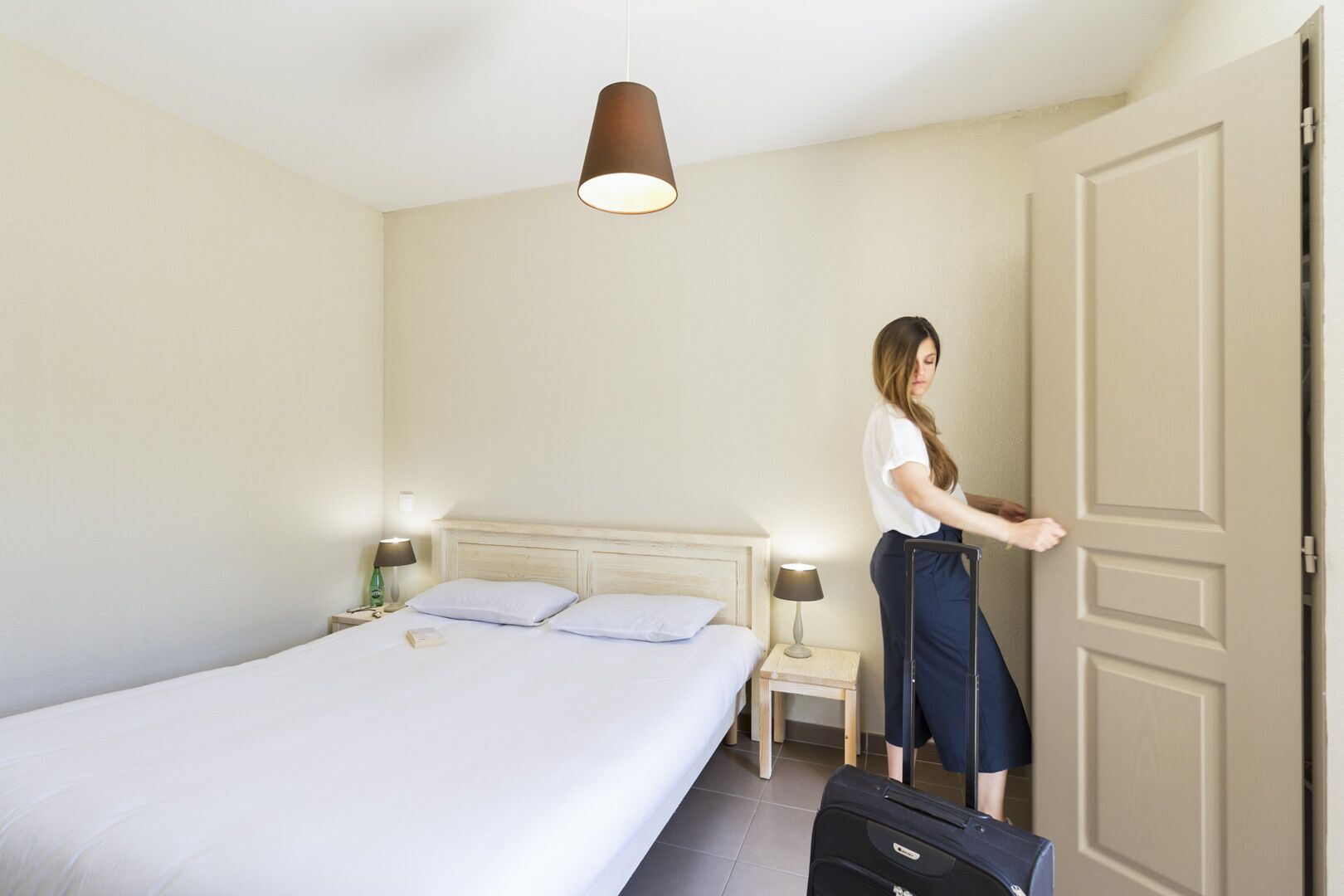 A hotel room with a bed, bedside tables, a lamp, a suitcase, and a door.