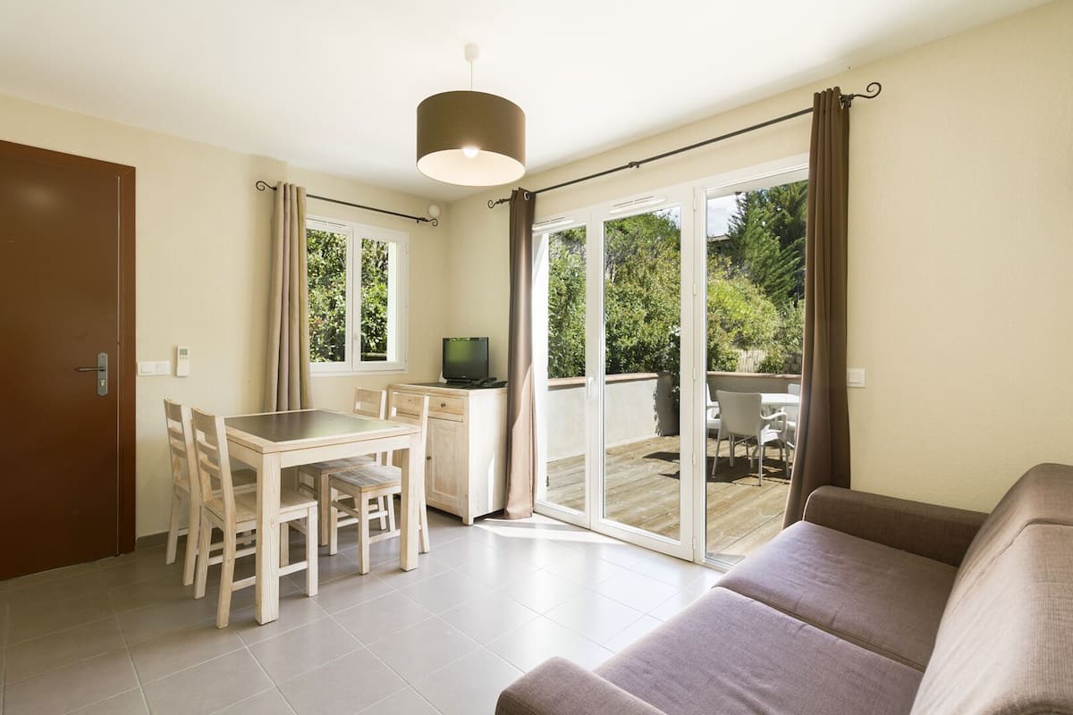 A modern living room with a sofa, a dining table with chairs, a TV, and a sliding glass door leading to an outdoor patio.