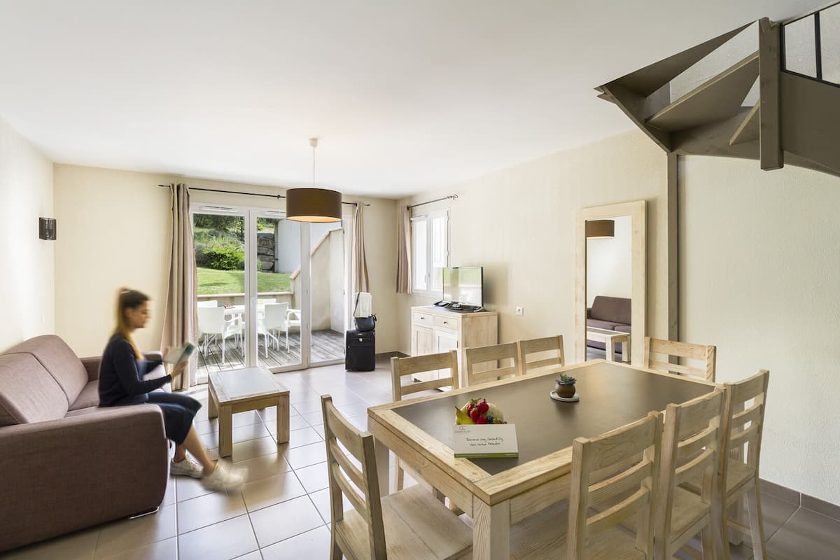 A modern living room with a sofa, a dining table with chairs, a laptop on a desk, and a view of the outdoors through a sliding door.