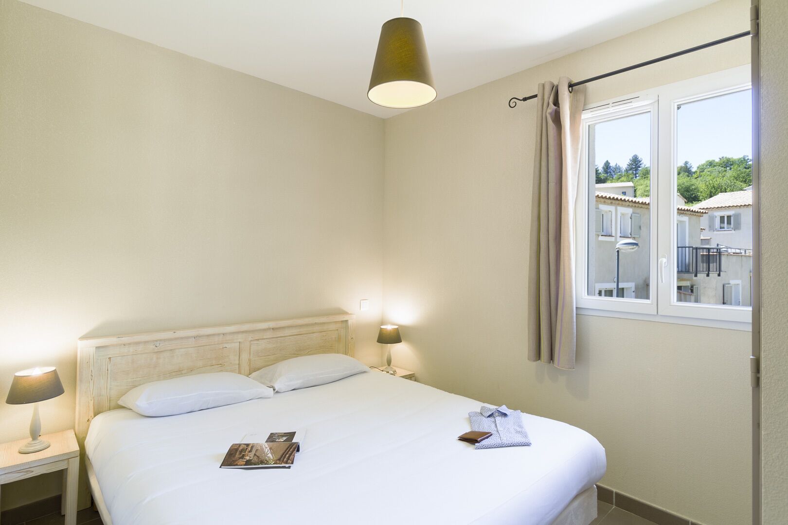 Une chambre d’hôtel avec un lit, deux lampes de chevet, une fenêtre avec des rideaux et une vue sur les bâtiments environnants.