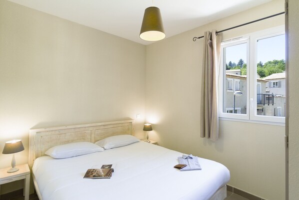 A hotel room with a bed, two bedside lamps, a window with curtains, and a view of buildings outside.