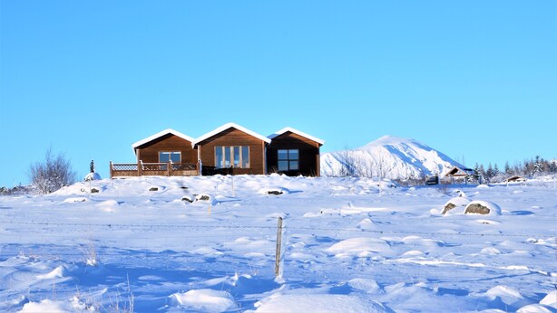 A wooden cabin with a large window and a door, surrounded by snow-covered ground and a clear blue sky. - Uthlid Cottages (Bláskógabyggd)