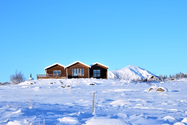 A wooden cabin with a large window and a door, surrounded by snow-covered ground and a clear blue sky. - Uthlid Cottages (Bláskógabyggd)