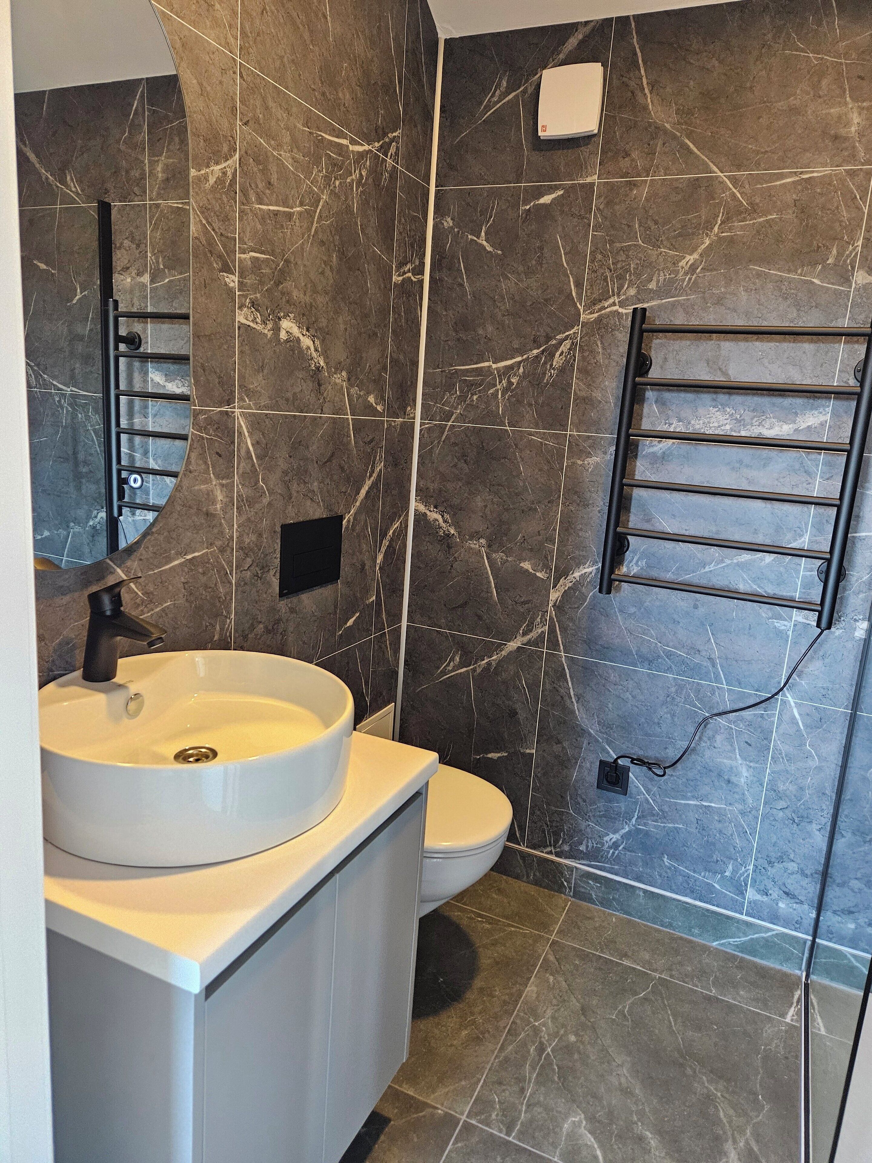 Een moderne badkamer met een witte wastafel, een zwarte kraan en een toilet. De wanden zijn betegeld met donkere marmertegels met witte aders.
