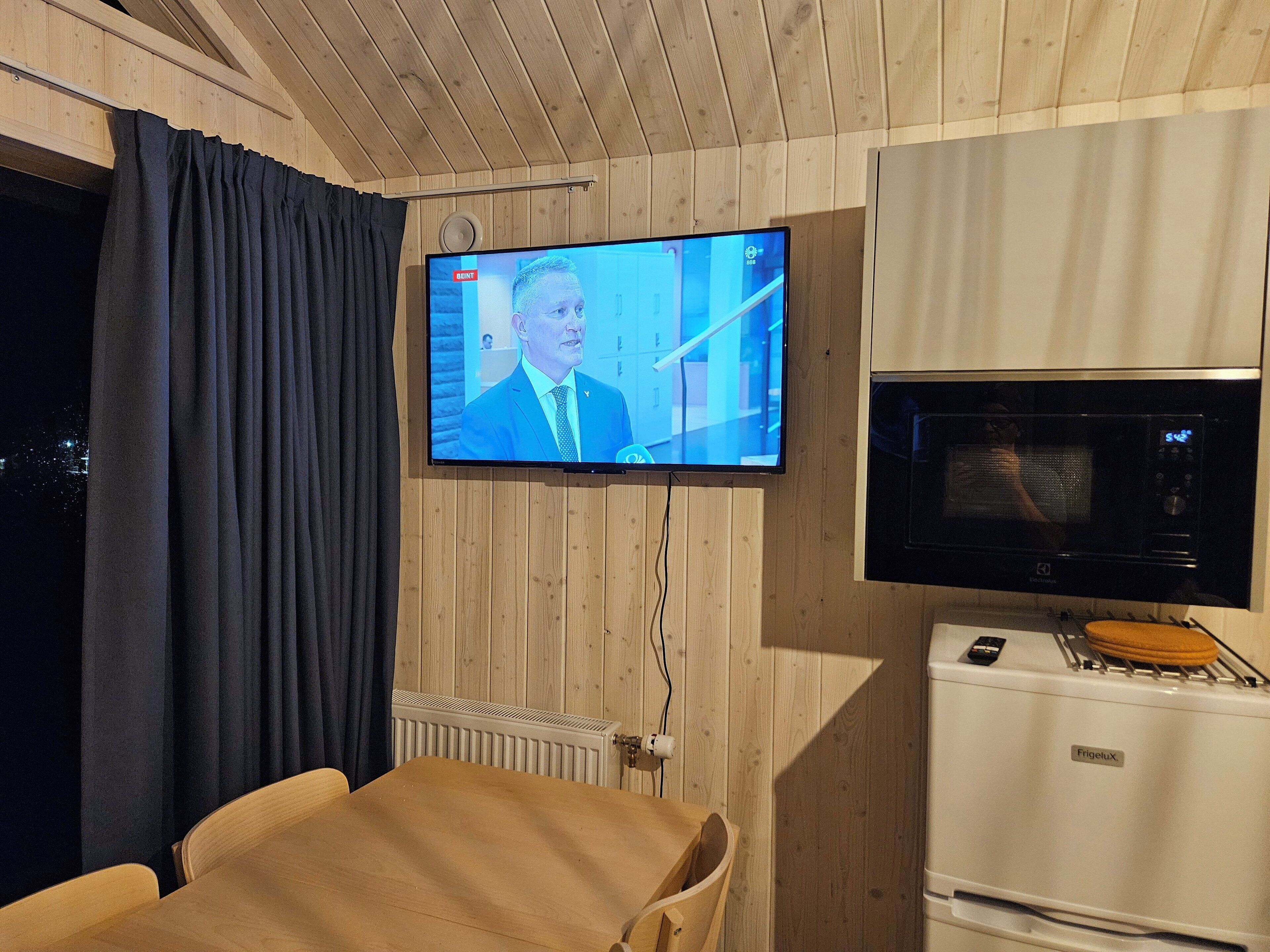 A room with a television mounted on the wall, a microwave, and a wooden ceiling.
