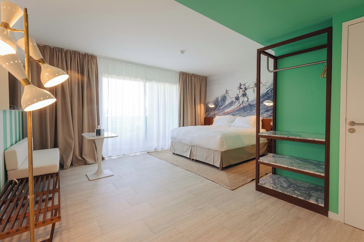 Une chambre d’hôtel comprenant un lit, un coin salon avec un fauteuil, une petite table, une étagère avec des objets décoratifs et un mur d’accent de couleur verte.