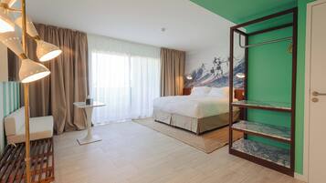 Habitación de hotel con cama, zona de estar con una silla, una mesita, estante con objetos decorativos y pared verde.