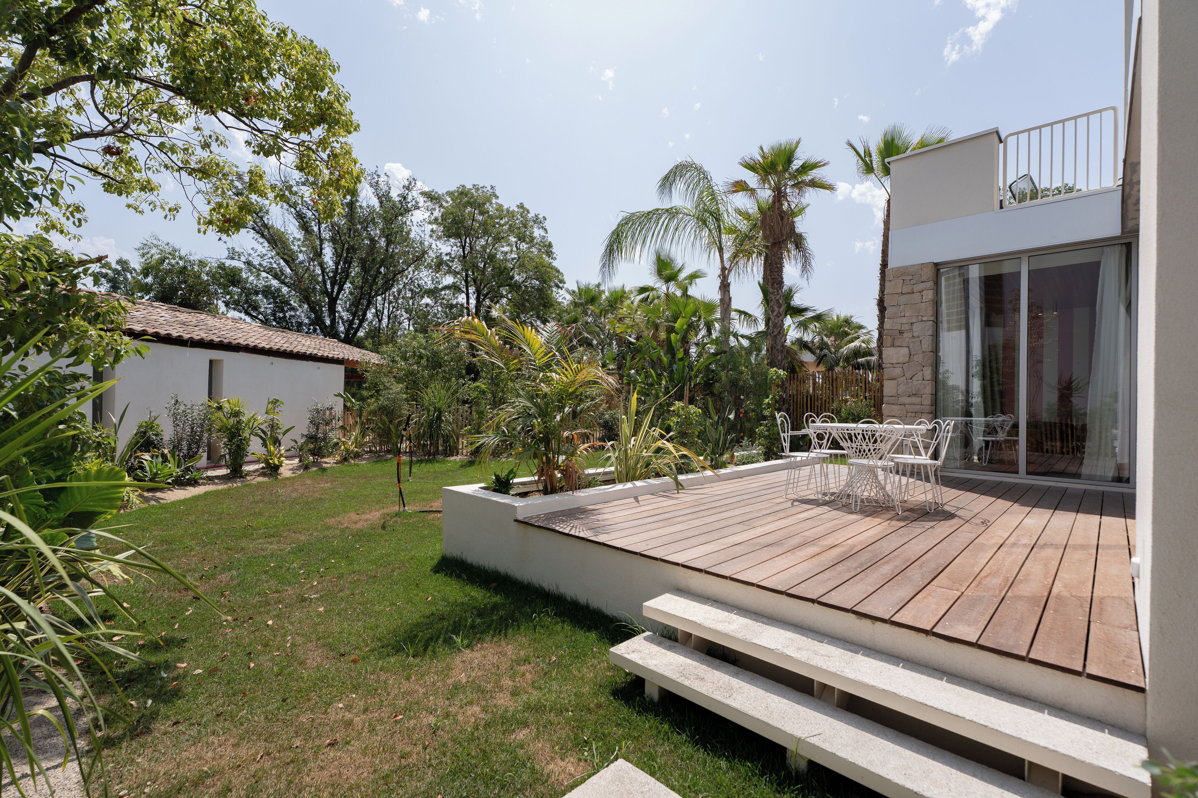 Eine moderne Terrassenanlage im Freien mit Holzboden und weißen Bänken, umgeben von üppiger Vegetation und Palmen.