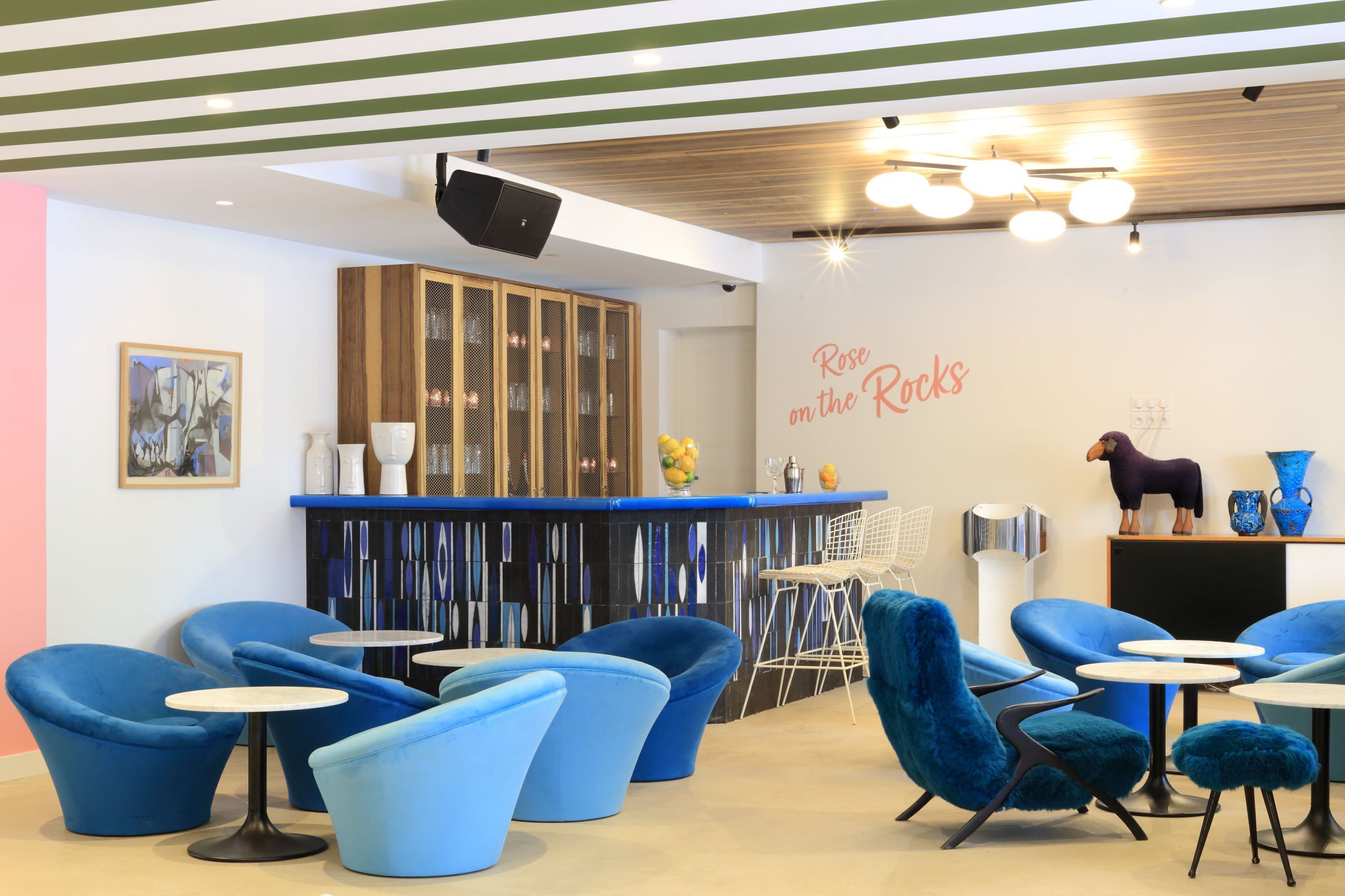 Un área de estar moderna con sillas y mesas azules, una barra con un fondo decorativo y una pared con el texto "Rose on the Rocks".
