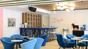 Un área de estar moderna con sillas y mesas azules, una barra con un fondo decorativo y una pared con el texto "Rose on the Rocks".