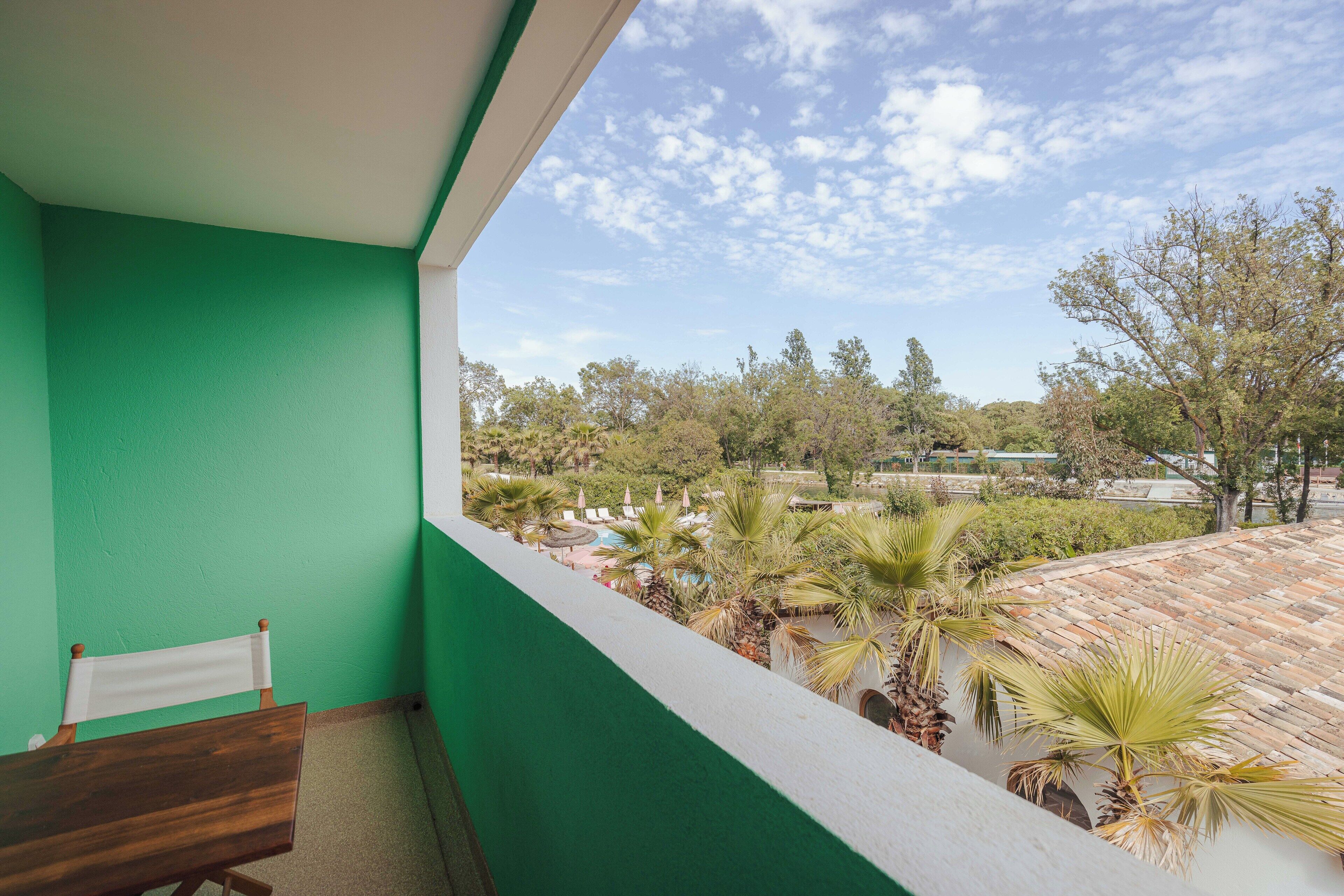 A balcony with a view of a pool and palm trees, a wooden chair, and a green wall.