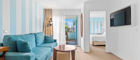 A modern hotel room with a blue sofa, a round wooden table, a TV mounted on the wall, and a view of the outside balcony.