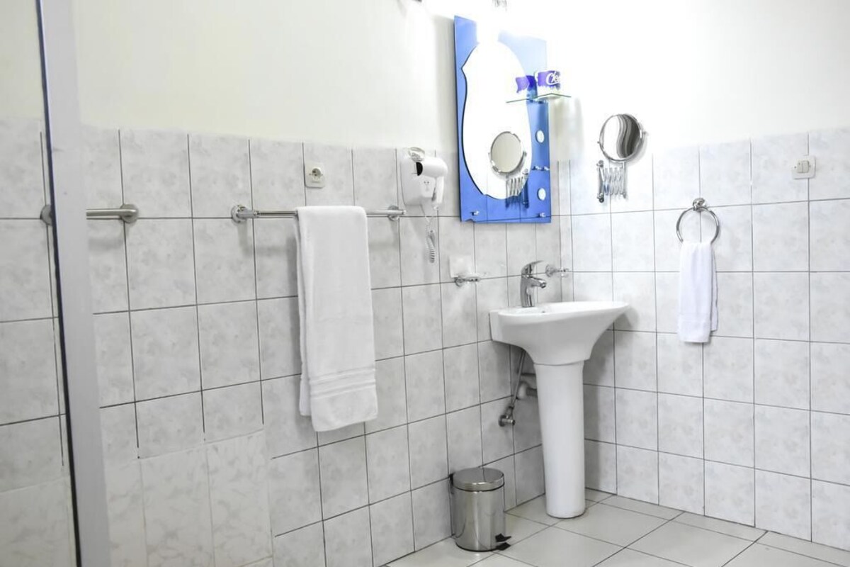 A bathroom with white tiles, a sink, a mirror, a towel rack, and a trash can.