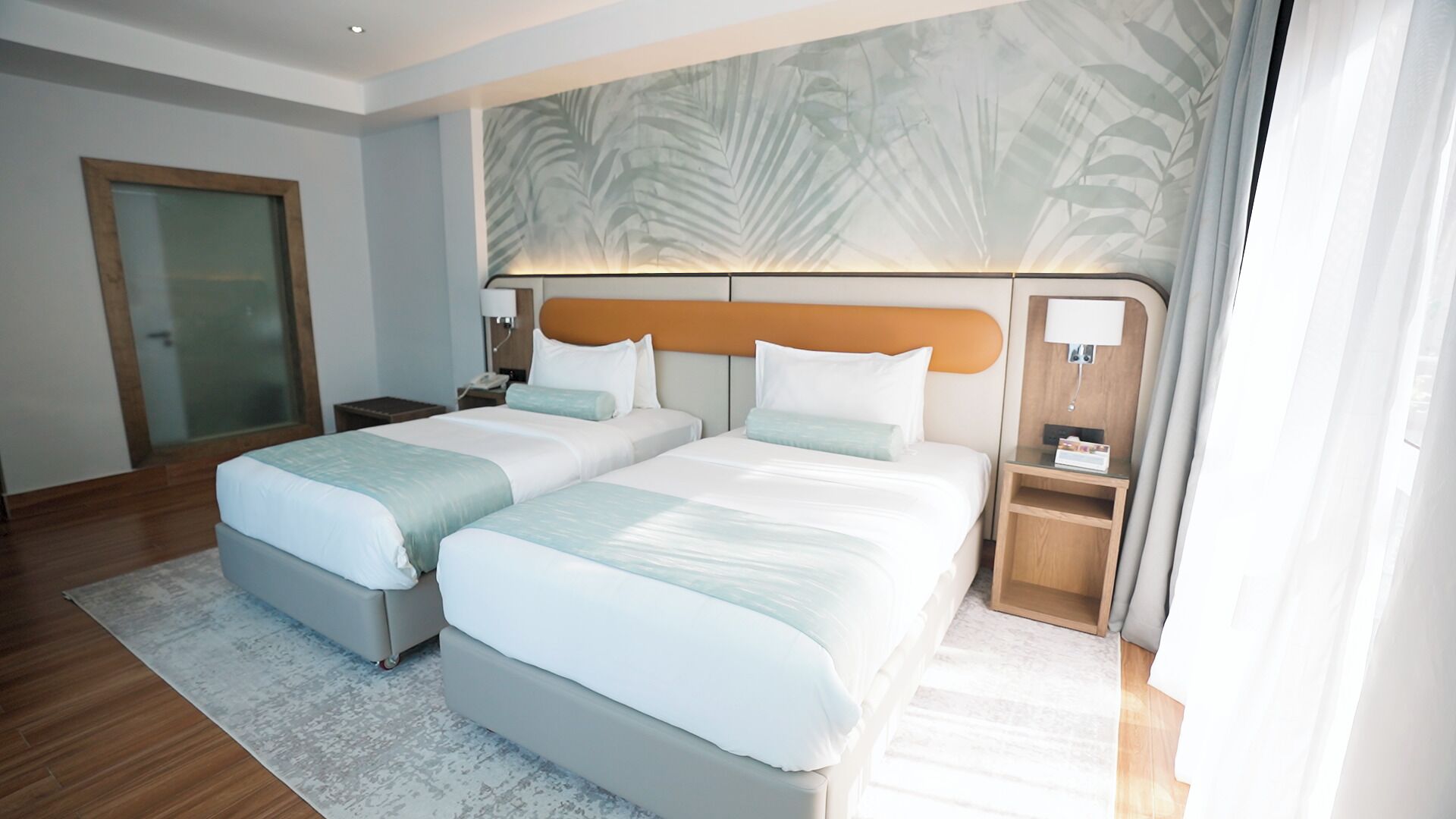 Habitación de hotel con dos camas, un ventanal con cortinas, un cabecero de madera y una lámpara de pared.