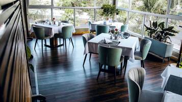 Un restaurant doté de nombreuses tables et chaises, de grandes fenêtres et de plantes d’intérieur.
