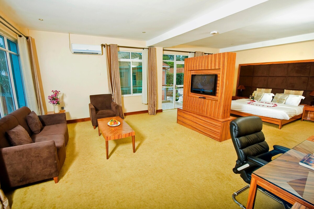 A hotel room with a sofa, armchair, TV, bed, and a small table with a flower vase.