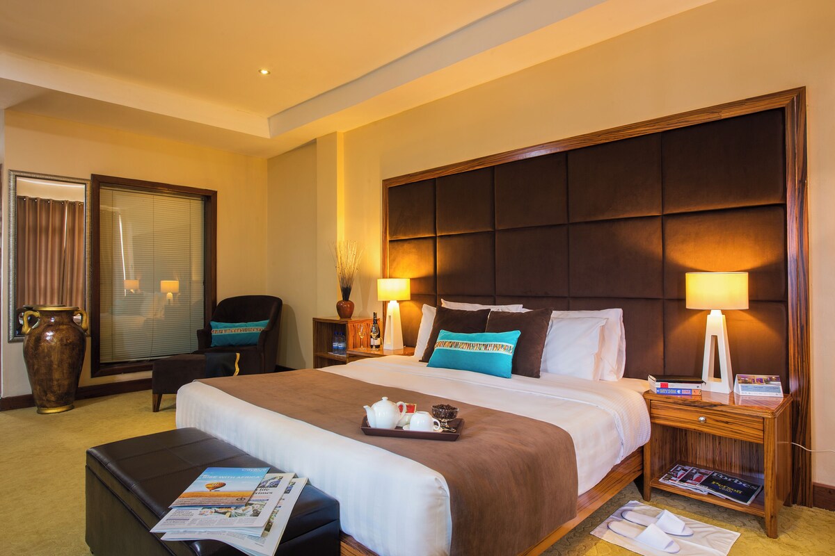 A hotel room with a large bed, a nightstand with a lamp, a chair, a large vase, and a window with curtains.