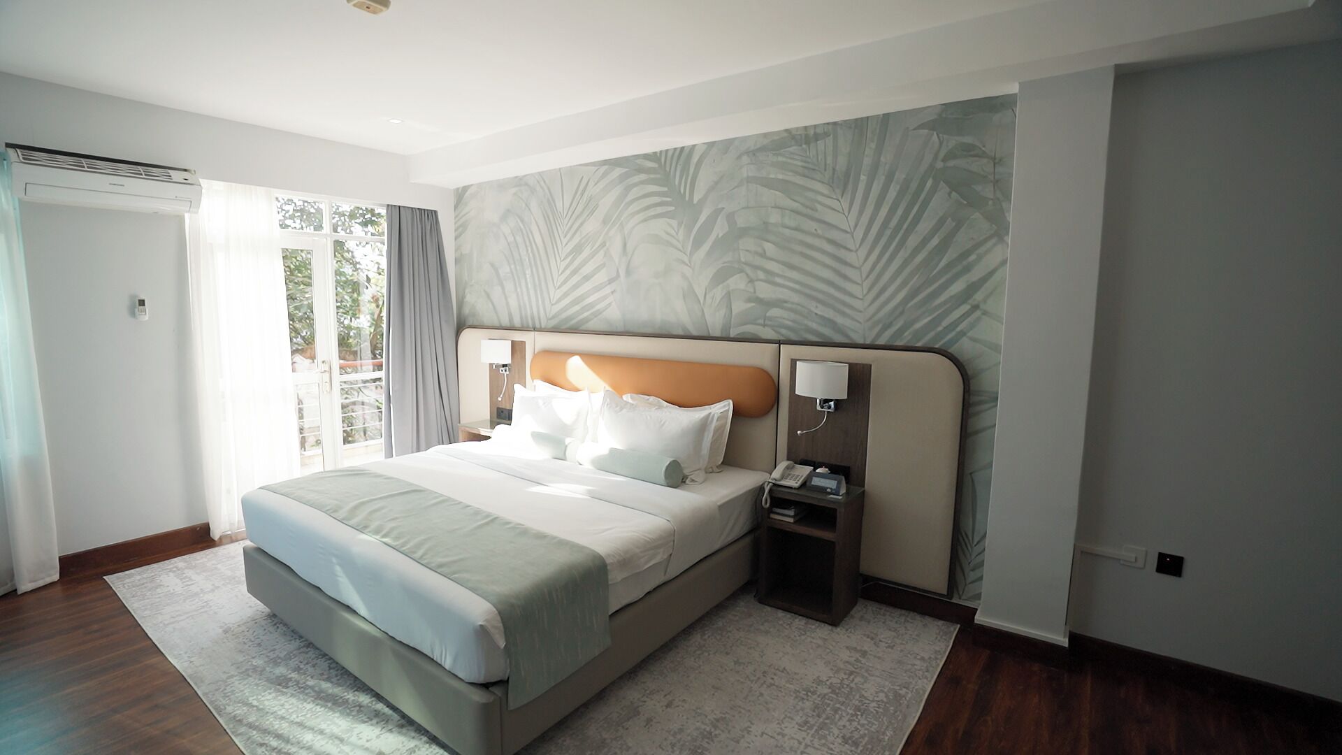 A hotel room with a large bed, a bedside table with a lamp, a telephone, and a view of the outdoors through a sliding door.
