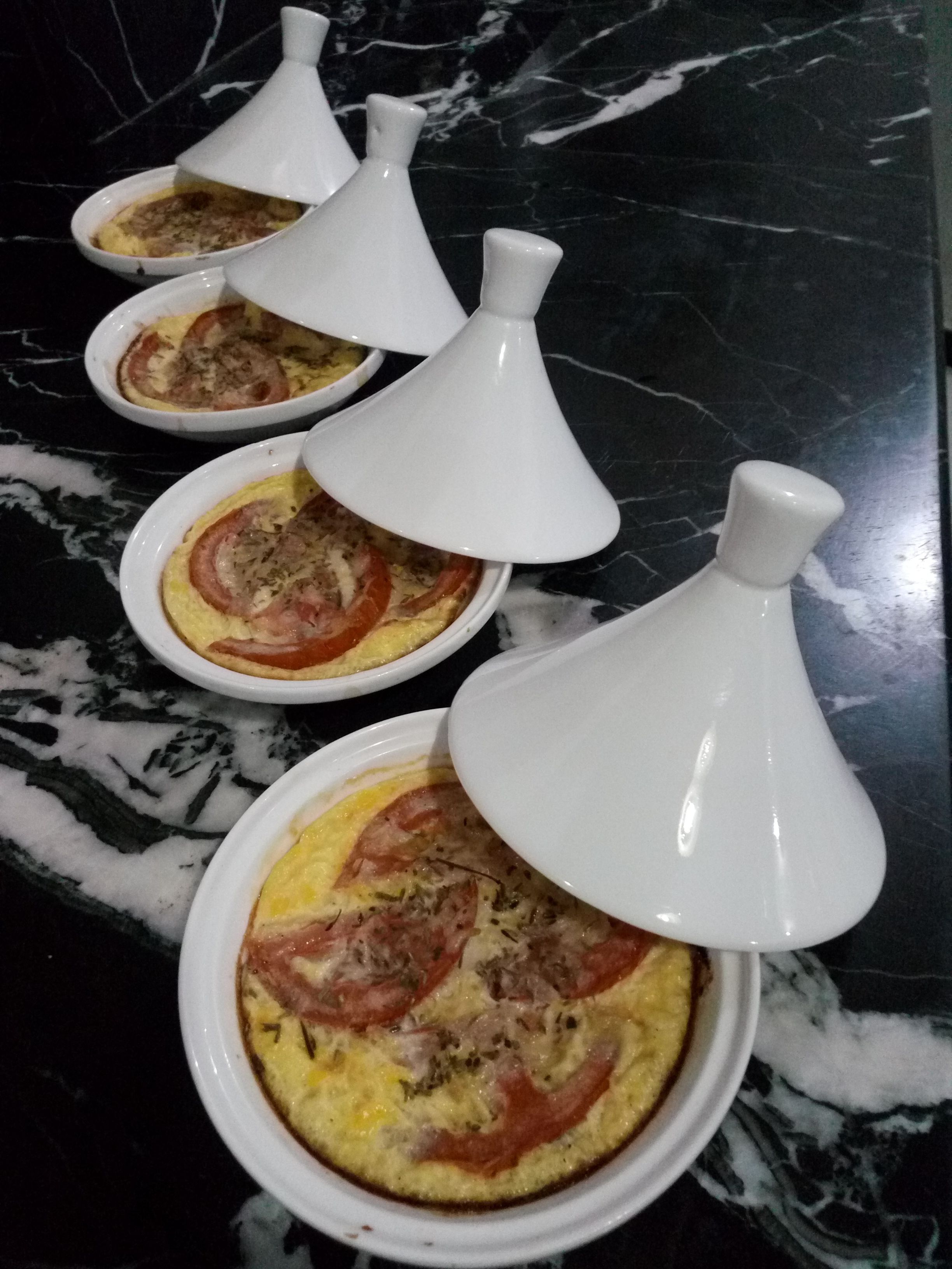Cuatro recipientes blancos de cerámica con tapas, que contienen comida, sobre una encimera de mármol oscuro.