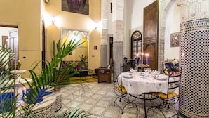 Un interior tradicional marroquí con comedor que presenta una mesa preparada para dos personas, rodeado de decoración elaborada y suelo de azulejos.