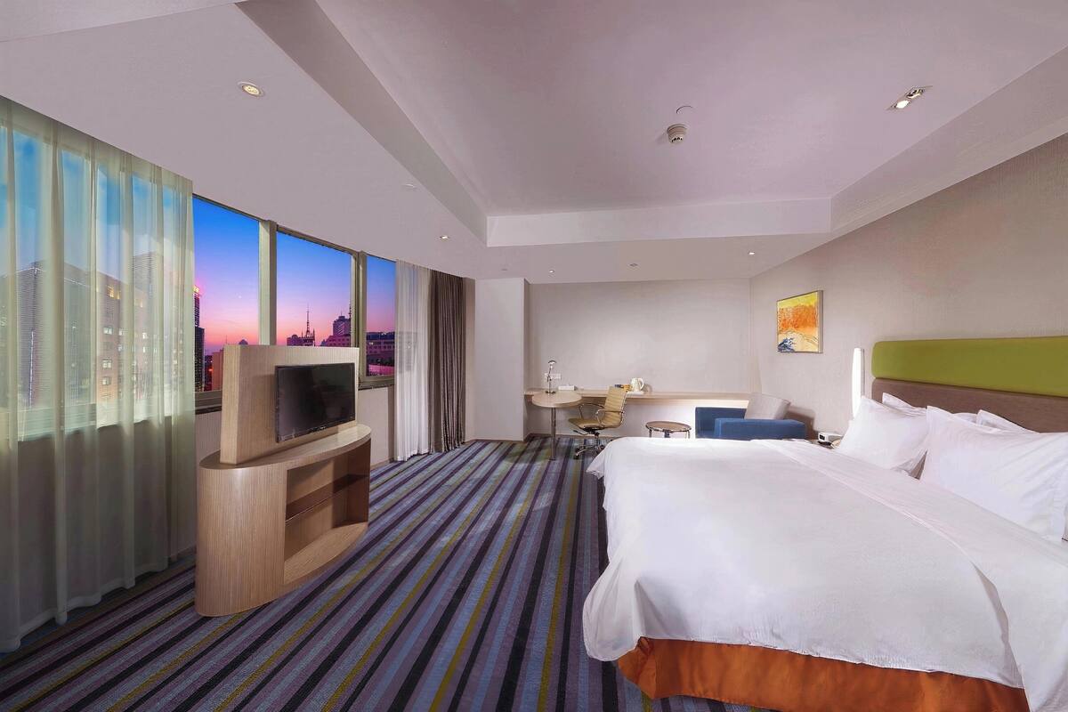 A hotel room with a large bed, a TV on a wooden stand, a desk with a chair, and a view of a cityscape through large windows.