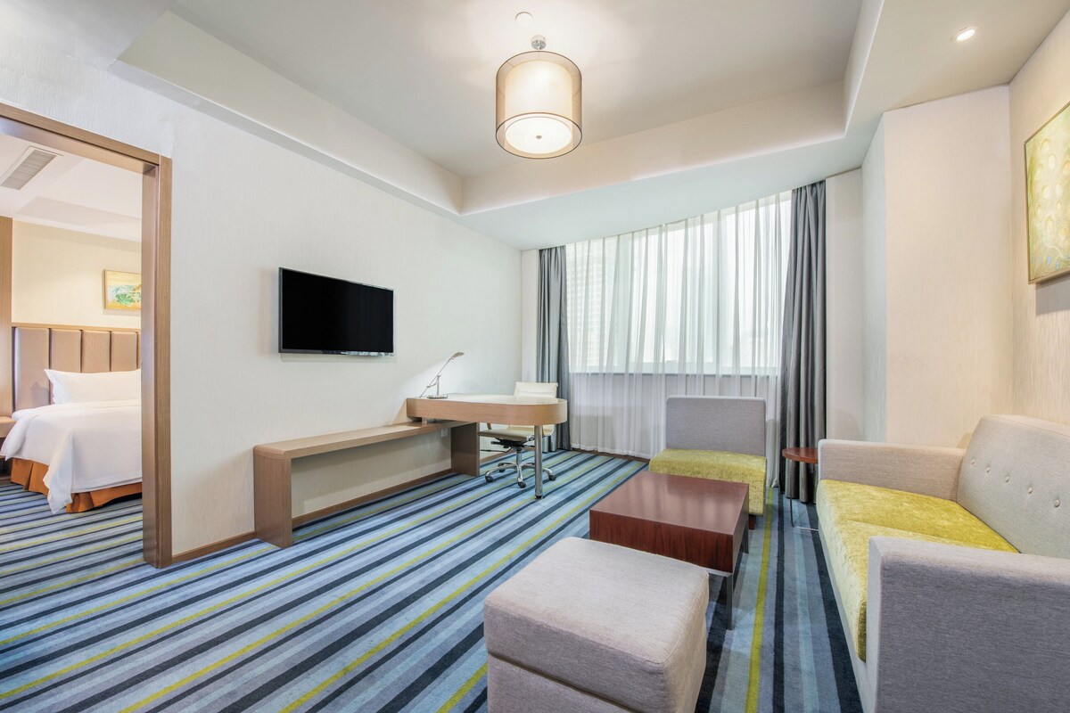 Habitación de hotel con cama, escritorio, silla, sofá, televisor y un amplio ventanal con cortinas.