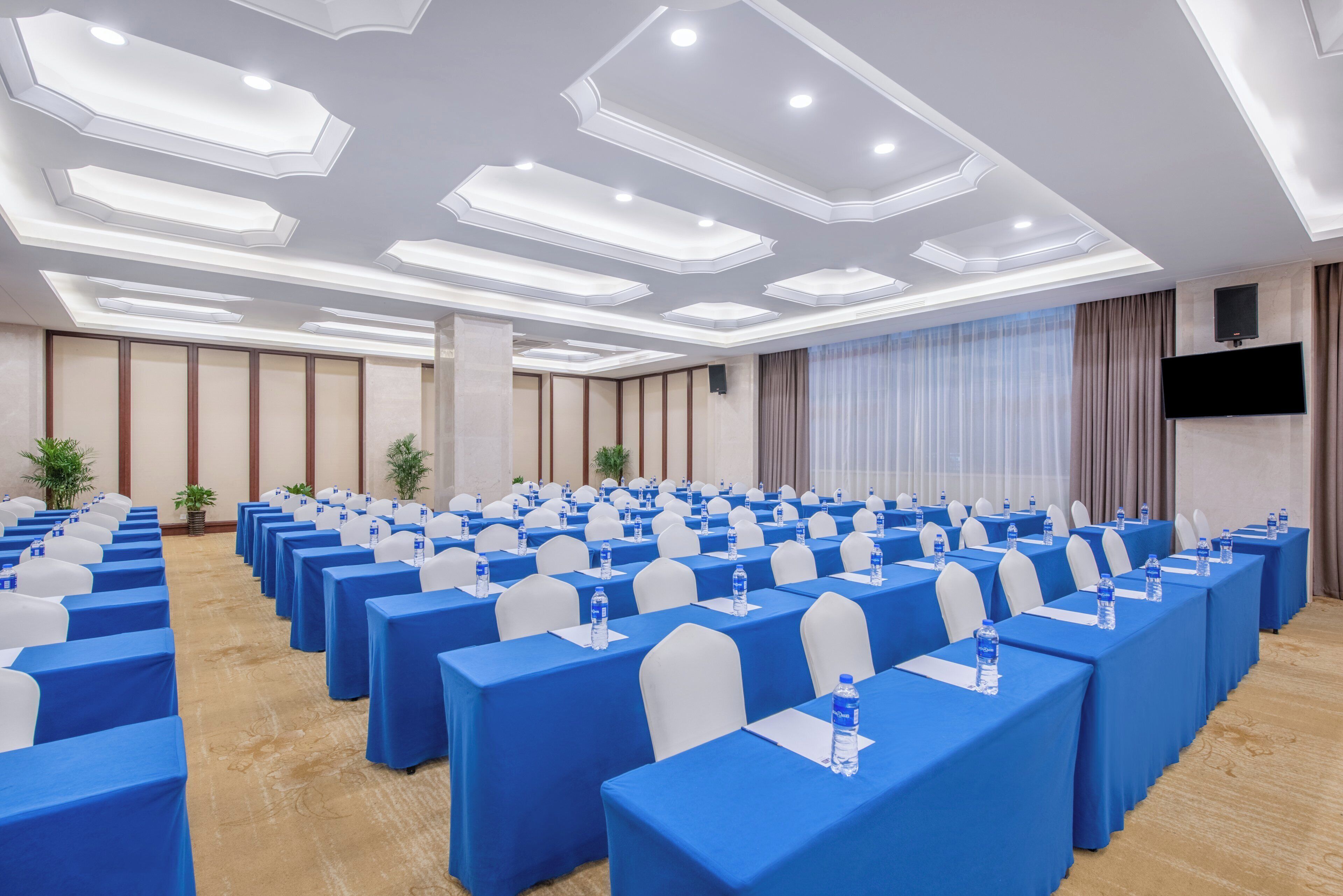 A conference room with rows of tables, chairs, and water bottles, featuring a modern ceiling design and a flat-screen TV.