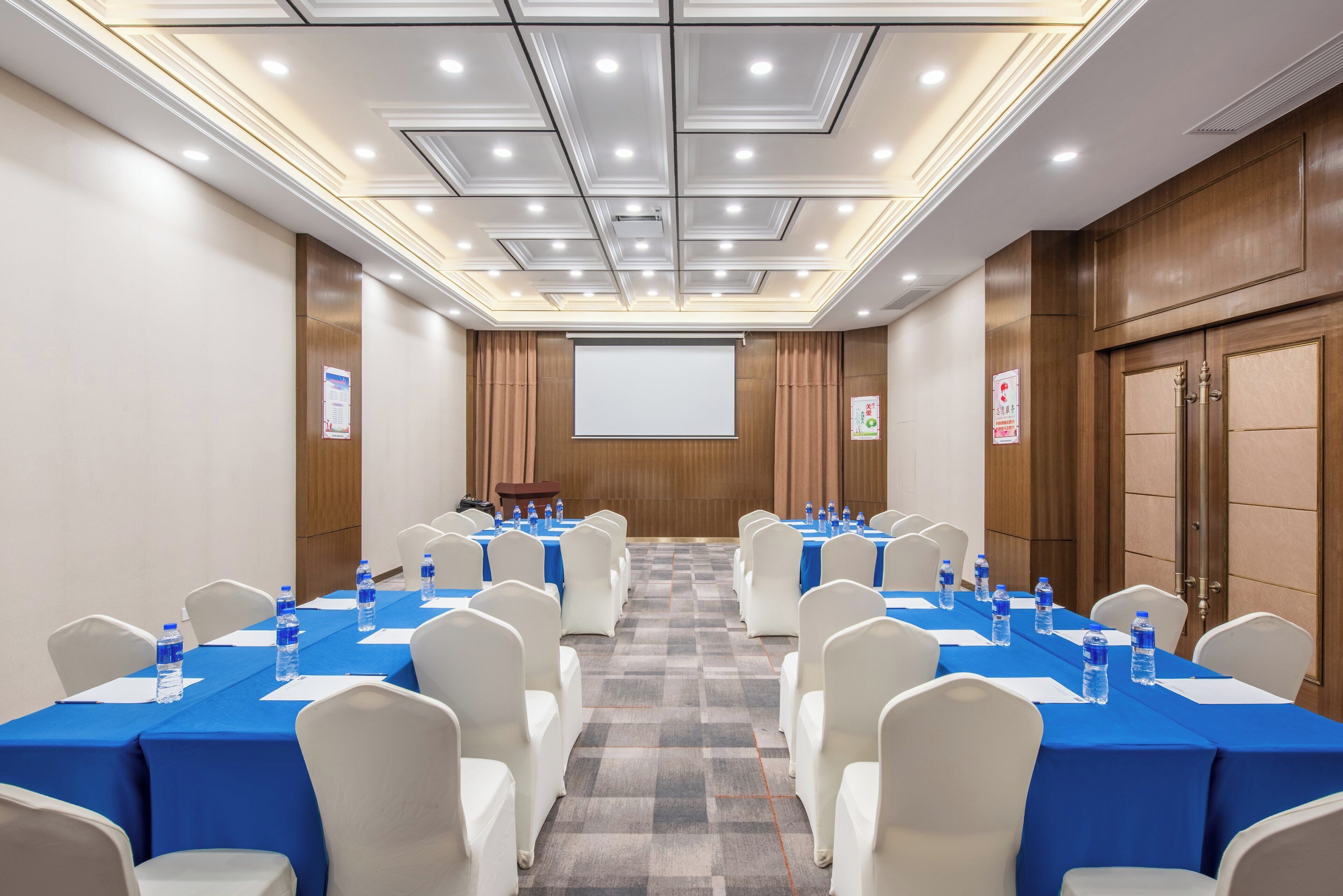 A conference room with tables set for meetings, equipped with water bottles, and a projector screen at the front.