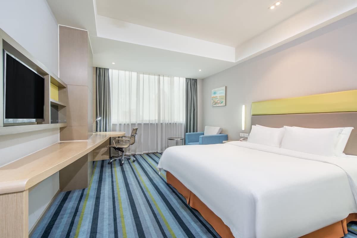 A hotel room with a large bed, a desk, a chair, a TV, and a window with curtains.