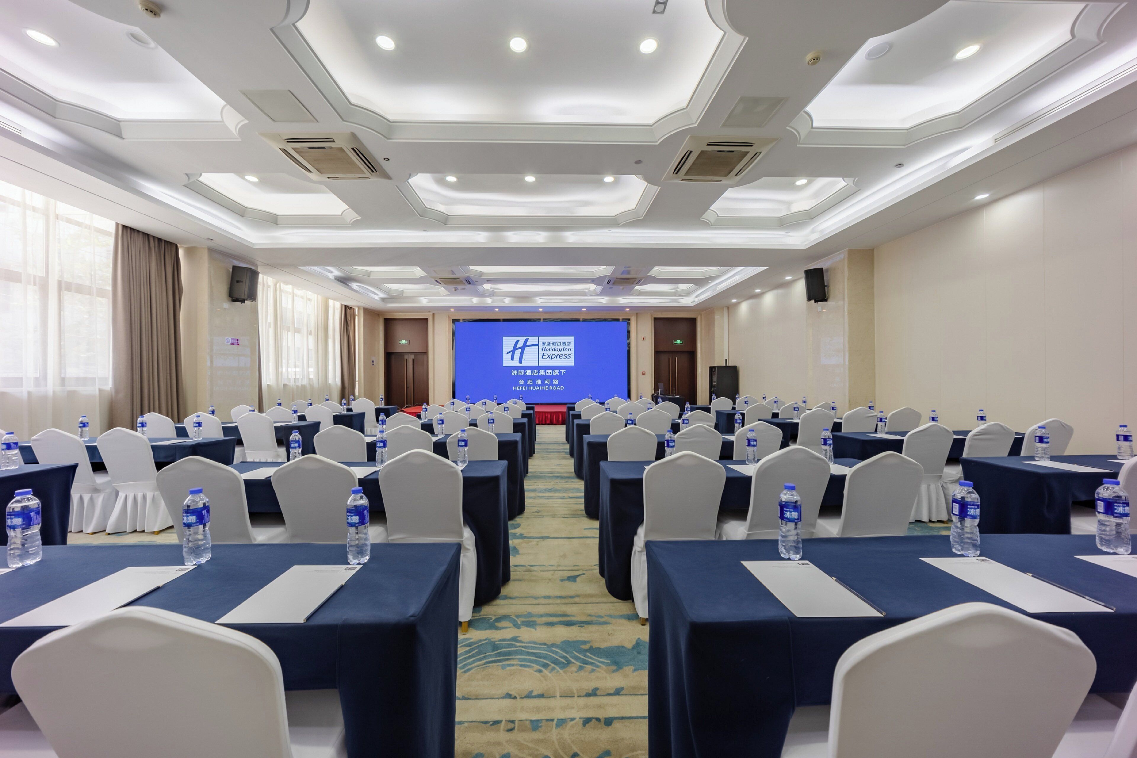A conference room with rows of tables, chairs, and a large screen at the front.