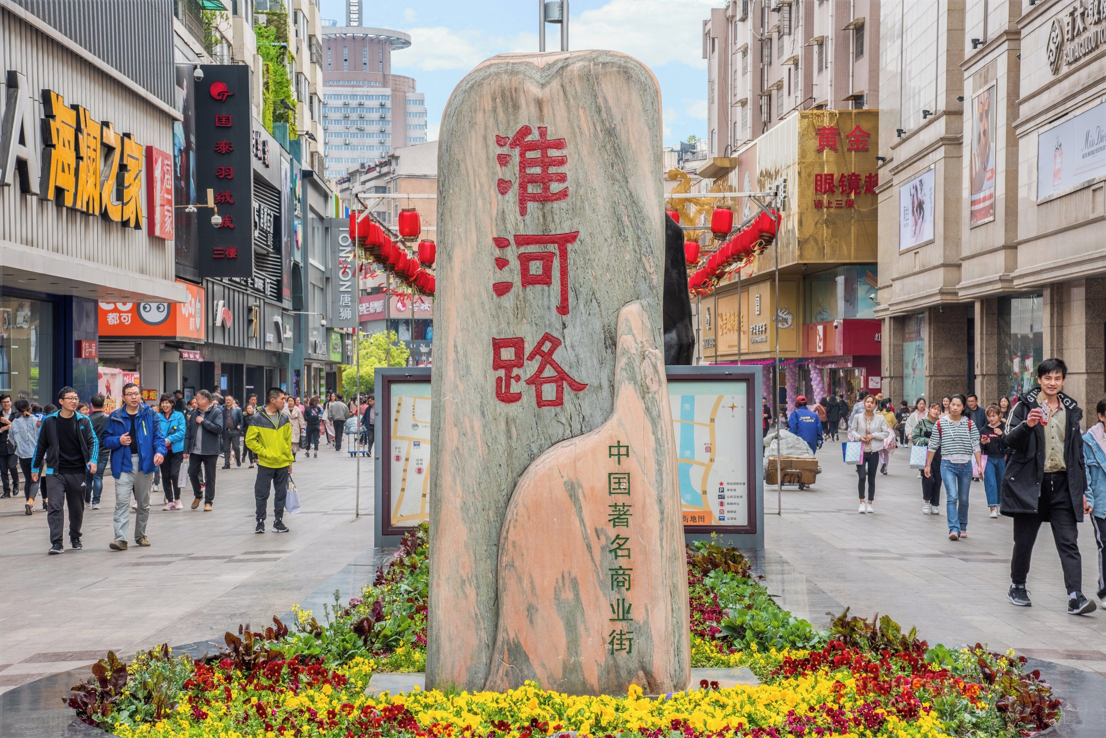 A stone monument with inscriptions in a public square surrounded by flower beds and busy pedestrian traffic.