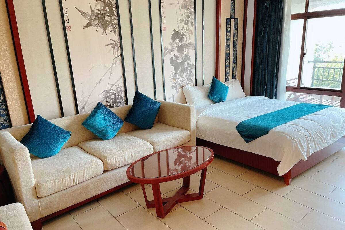A hotel room with a sofa and a bed, both with blue pillows. There is a wooden table in the center.