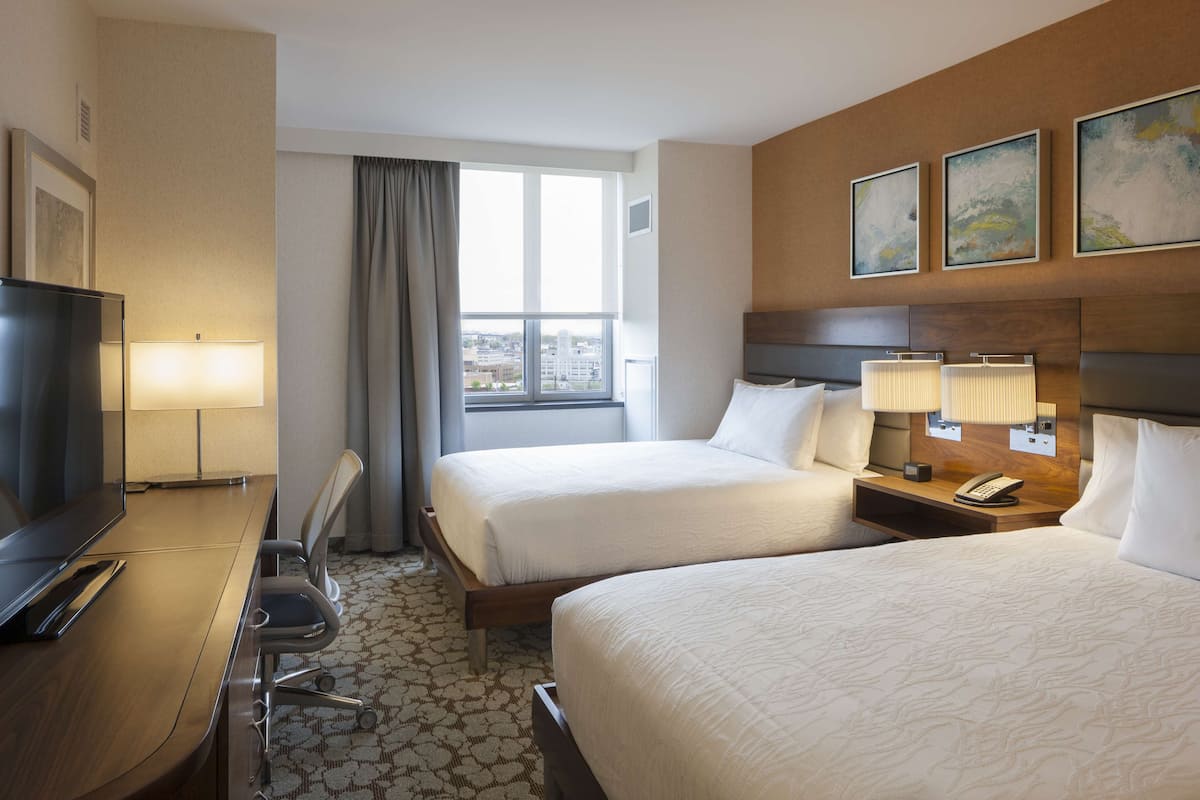 A hotel room with two beds, a desk with a TV, a chair, a window with curtains, and artwork on the wall.