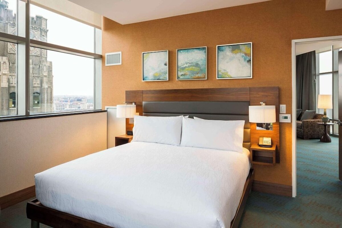 Habitación de hotel con una cama grande, dos mesitas de noche con lámparas, vista a un paisaje urbano y tres cuadros enmarcados en la pared.