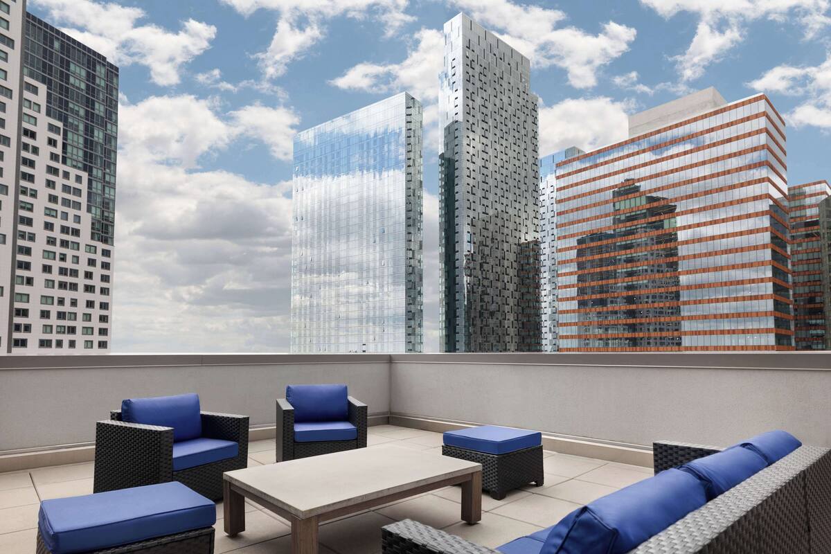 A rooftop terrace with wicker furniture, a table, and a cityscape view of modern buildings.