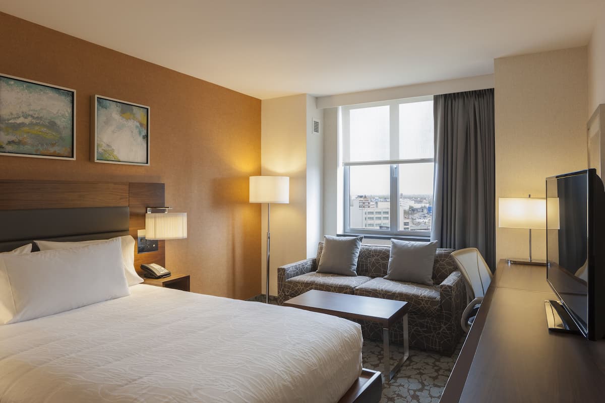 A hotel room with a large bed, a sofa, a desk, and a television. There are two paintings on the wall and a window with curtains.