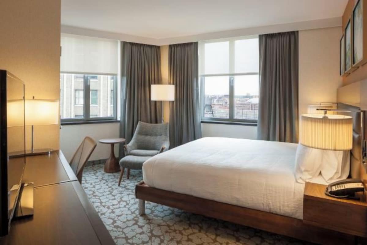 Habitación de hotel con cama, escritorio, silla, lámpara y vista de la ciudad a través de amplias ventanas.