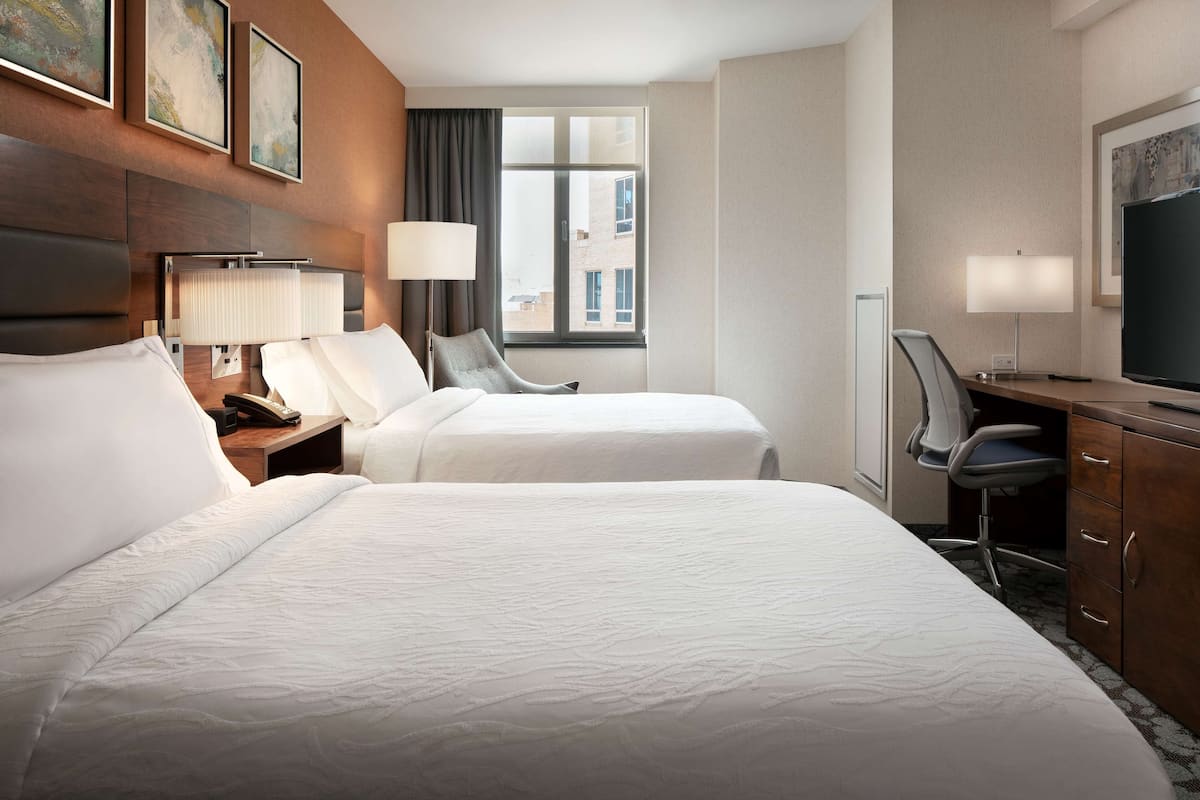 A hotel room with two beds, a desk, a chair, a TV, and artwork on the walls.