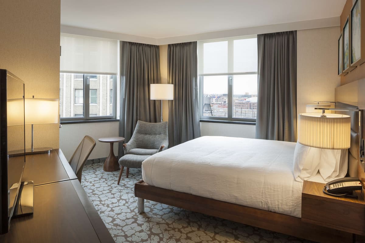 A hotel room with a large bed, a desk, a chair, a small table, and two windows with curtains.