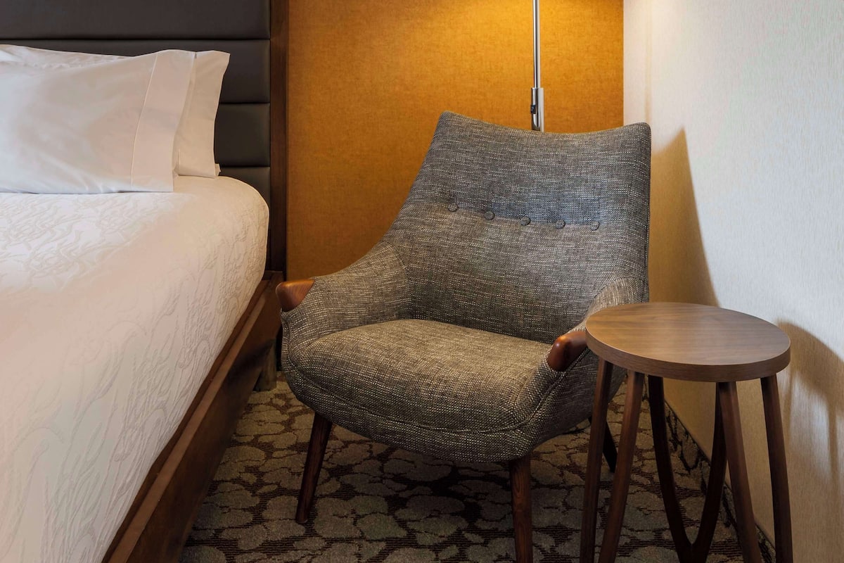 A hotel room with a bed, a grey armchair, a small wooden side table, and a floor lamp.