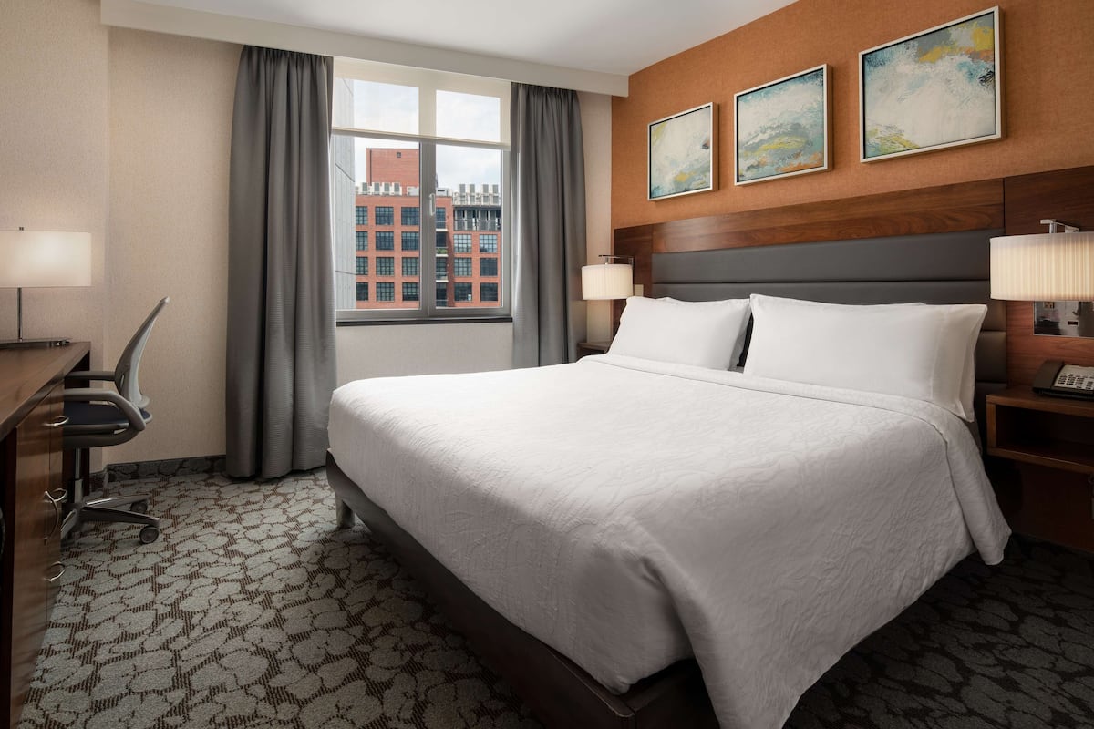 A hotel room with a large bed, a desk with a chair, a window with curtains, and artwork on the wall.