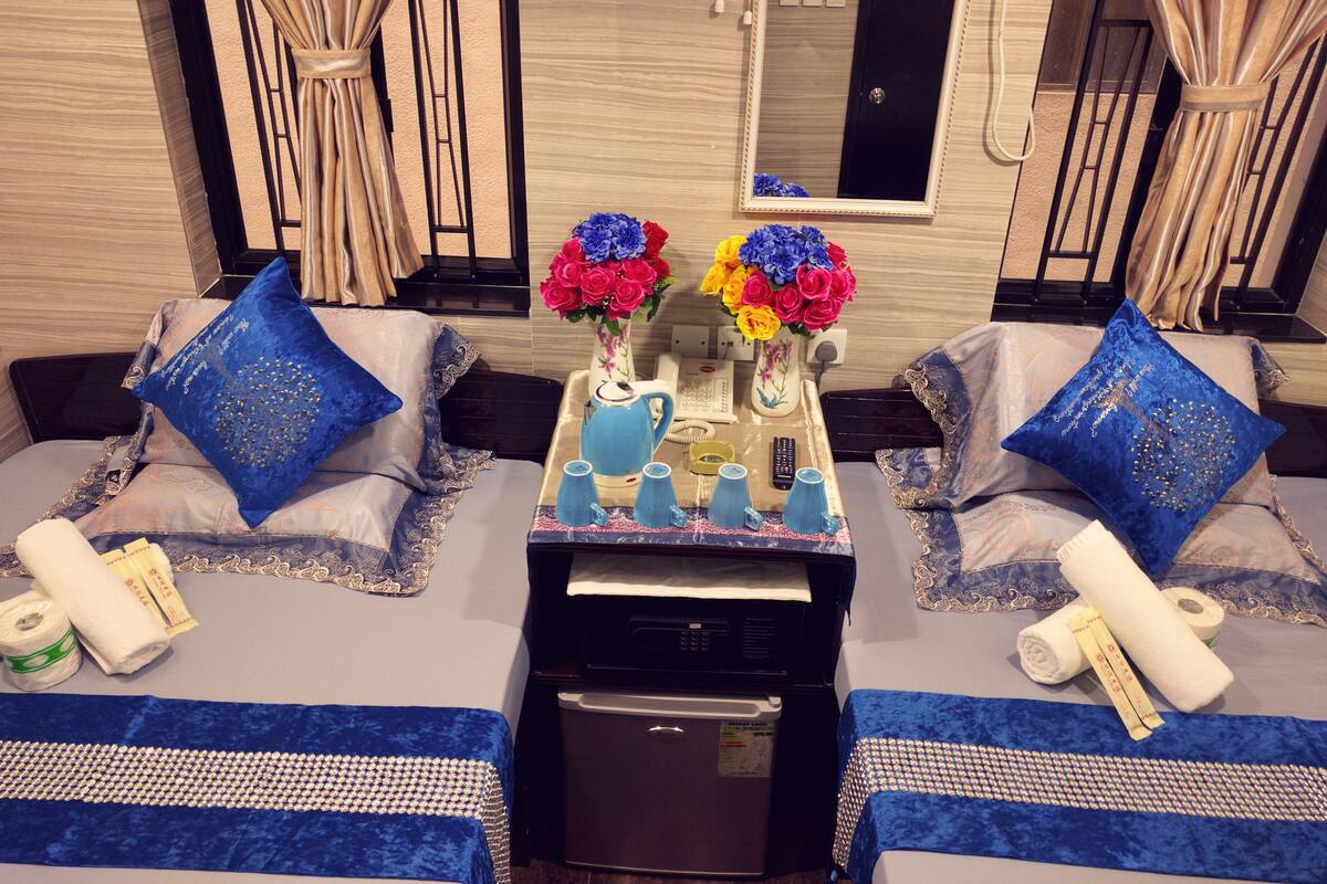 A hotel room with two beds, each with blue pillows and a blue bedspread. There are two vases with colorful flowers on a nightstand between the beds. A small refrigerator and a microwave are on the floor between the beds.