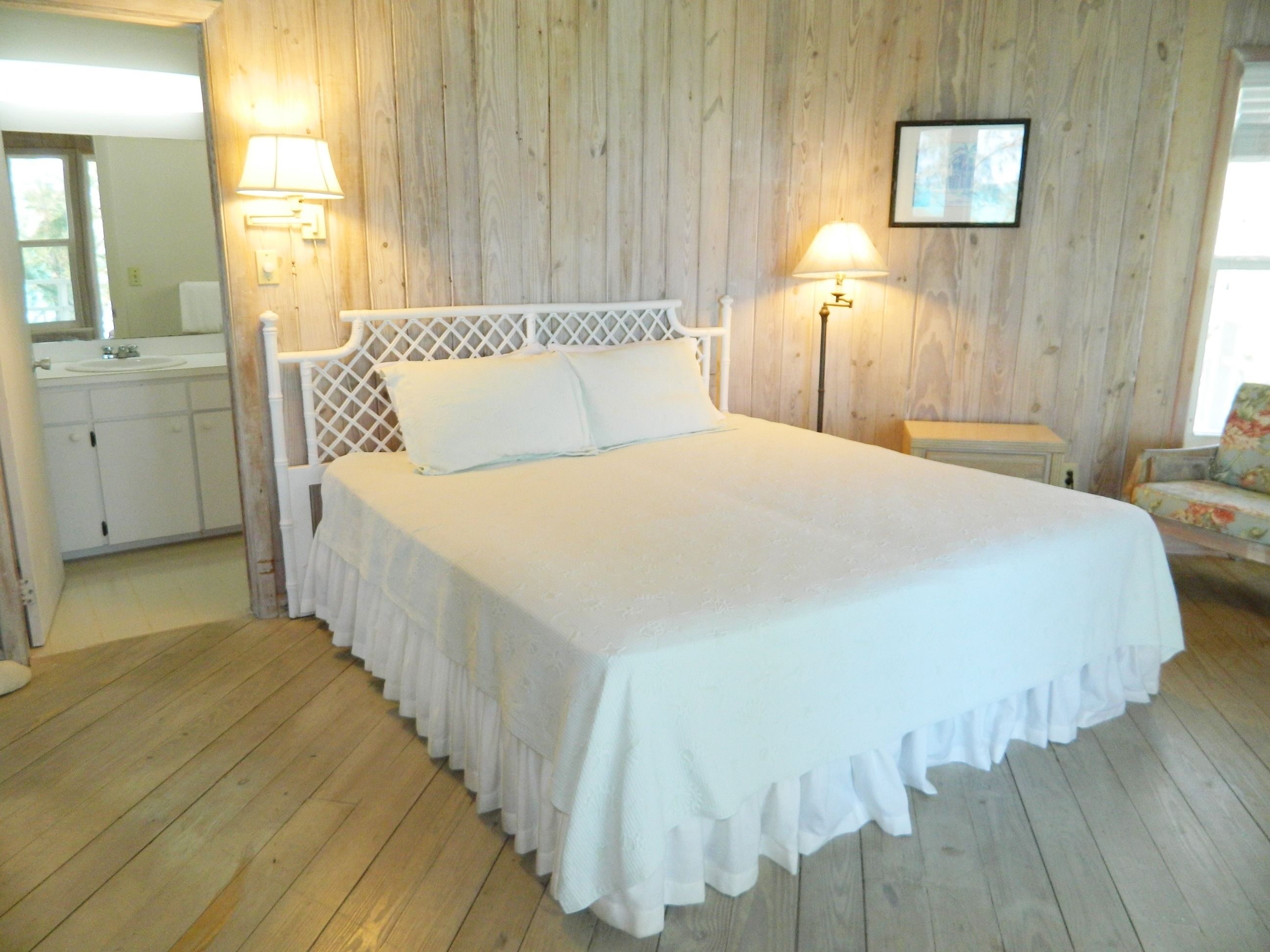 A bedroom with a wooden floor, a white bed with a ruffled bed skirt, two bedside lamps, a nightstand, a chair, and a framed picture on the wall.