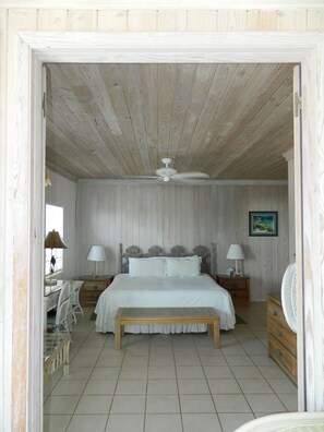A bedroom with a wooden ceiling, a bed with white bedding, two bedside tables with lamps, a wooden bench, and a fan. - Regatta Point (George Town)