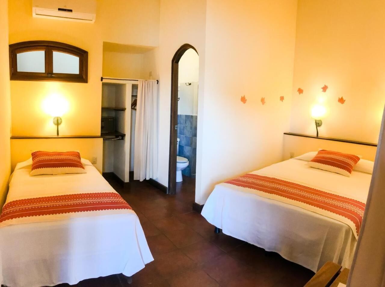 Ein Hotelzimmer mit zwei Betten, jeweils mit einer rot-weißen Bettdecke, einem Spiegel über dem linken Bett und einer Wand-Klimaanlage.