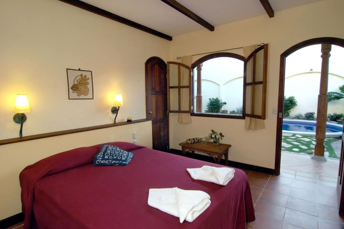 Ein Schlafzimmer mit einem großen Bett, zwei Nachttischlampen, einem kleinen Tisch mit einer Vase und Blick auf einen Swimmingpool durch ein Rundbogenfenster.