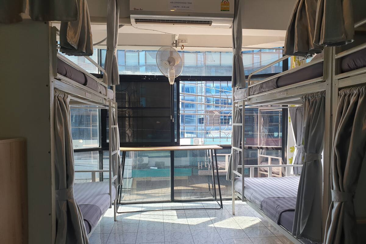 Habitación de albergue con literas, un ventilador y ventana con vista a un edificio.