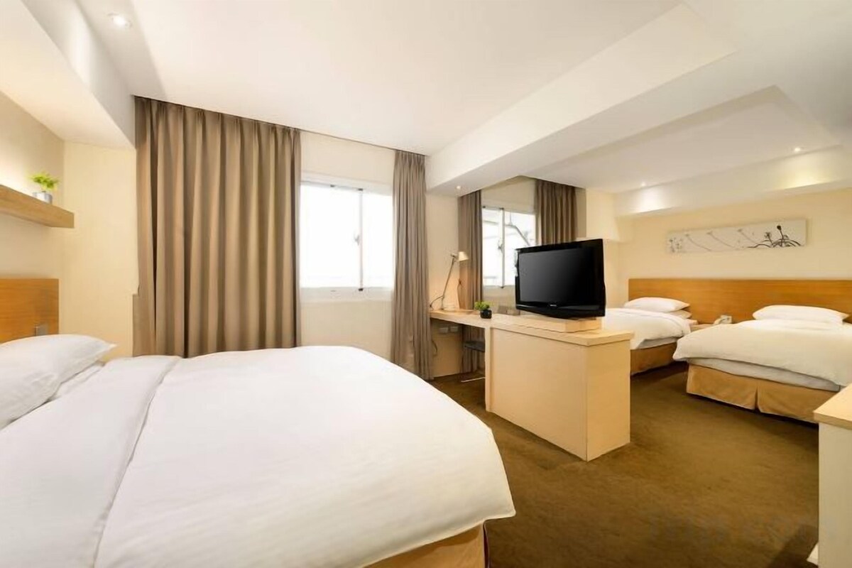 A hotel room with a large bed, a smaller bed, a desk with a TV, and a window with curtains.