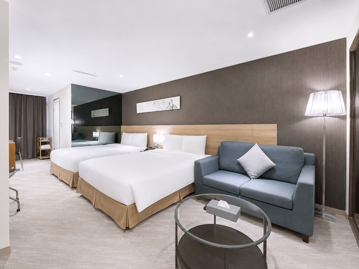 A hotel room with two beds, a sofa, a glass coffee table, and a lamp.