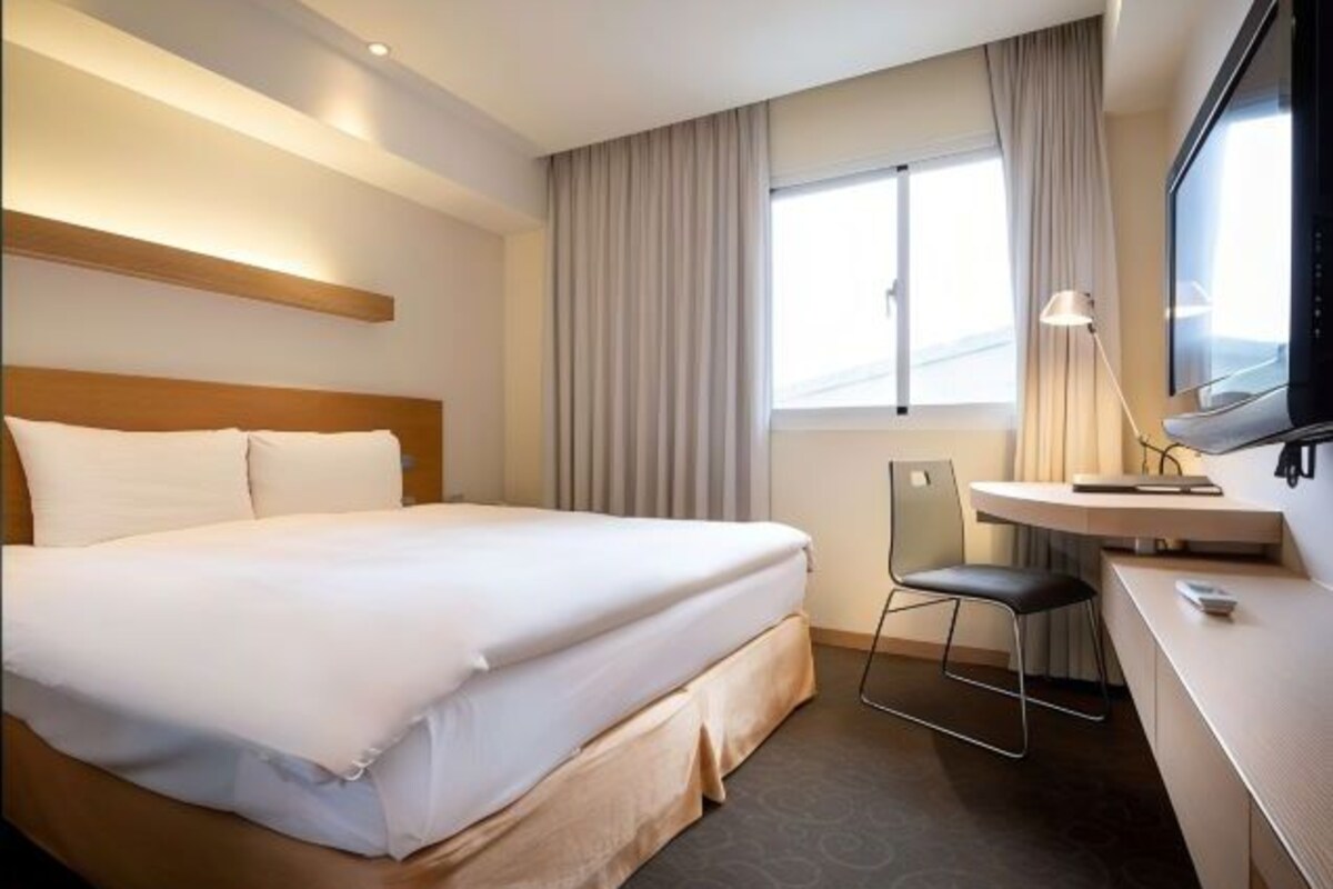 A hotel room with a large bed, a desk with a chair, a TV, and a window with curtains.